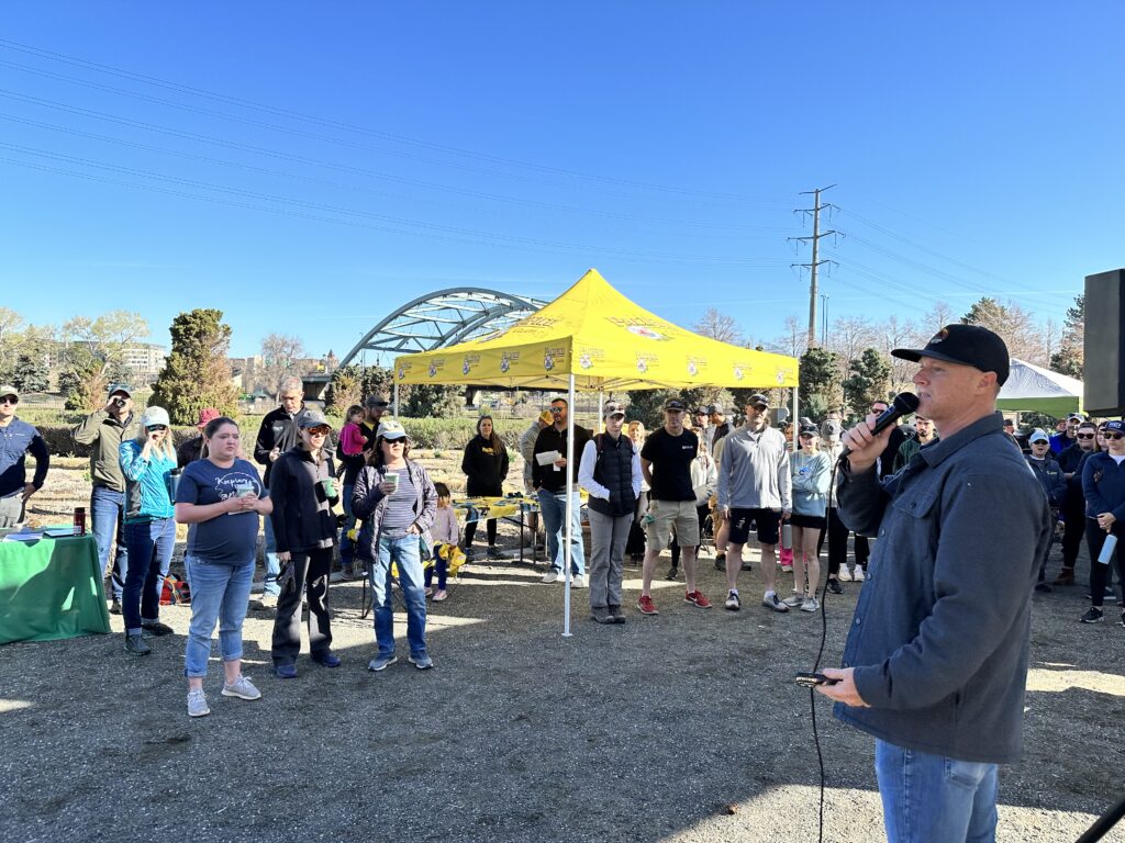 Ryan, TGF's Executive Director, wearing a grey shirt and black hat, speaking to a group of volunteers in the park ona  microphone. 
